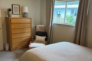 Une chambre à coucher avec un lit, une commode en bois, une chaise, une fenêtre avec des rideaux et une petite table.