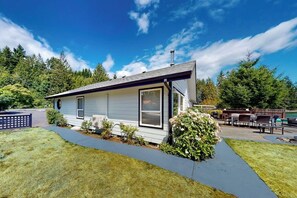 Une maison de plain-pied avec un porche à l’avant, une petite terrasse et un jardin à l’arrière équipé d’un barbecue et de meubles d’extérieur.