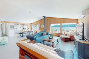 A spacious living area with a large window overlooking a lake, a cozy seating area, and a kitchen in the background. - Waterfront 3BR on Hood Canal with 3 kayaks & beach (Hoodsport)
