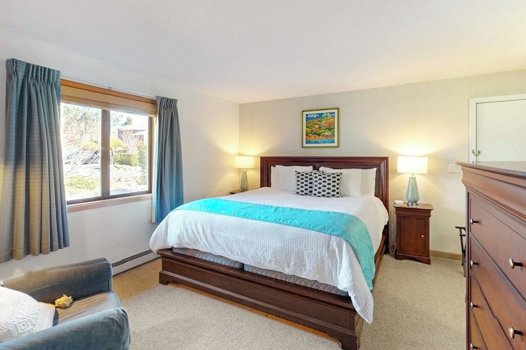 A hotel room with a bed, bedside lamps, a dresser, a chair, and a window with curtains.
