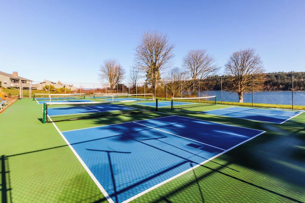 An outdoor tennis court with multiple courts, surrounded by trees and a body of water.