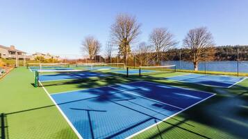 An outdoor tennis court with multiple courts, surrounded by trees and a body of water.