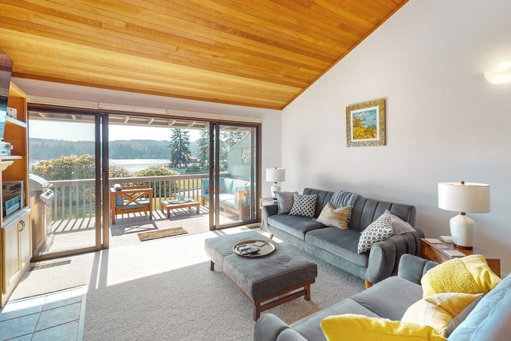 A modern living room with a grey sofa, a wooden ceiling, and a balcony with a view of trees and a lake.