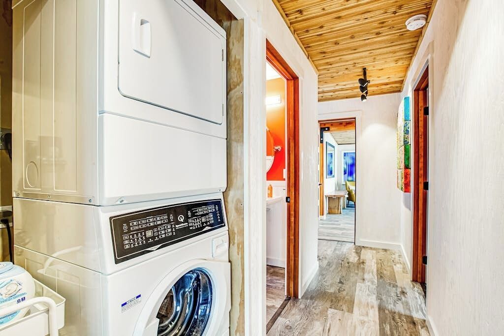 Une buanderie avec des machines à laver superposées, un plafond en bois et un couloir menant à une pièce dont un mur est rouge et une fenêtre est bleue.