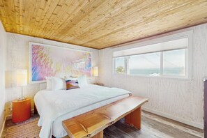 Ein Schlafzimmer mit Holzboden, einem großen Bett mit weißer Bettwäsche, einer Bank am Fußende des Bettes, einem farbenfrohen abstrakten Gemälde über dem Bett und einem Fenster mit Blick aufs Meer.