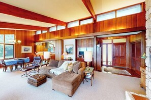 A spacious living room with a high ceiling, wooden paneling, a dining area with a blue tablecloth, and a fireplace. - Bayfront 1BR with patio, beach access, & views (Vashon)