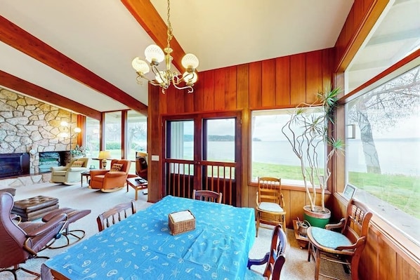 A room with a large table, chairs, a fireplace, and a view of the outdoors. - Bayfront 1BR with patio, beach access, & views (Vashon)