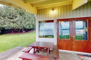 A wooden door with glass panels, a concrete patio with a wooden table, and a grassy yard with trees. - Bayfront 1BR with patio, beach access, & views (Vashon)