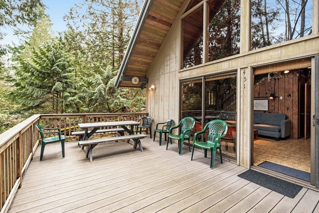 Una terraza de madera con mesas y sillas de picnic, una entrada con puerta de vidrio y vista a árboles.