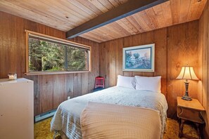 Ein Schlafzimmer einer Holzhütte mit einem Bett, einem Fenster, einem gerahmten Bild, einem Nachttisch und einer Lampe.