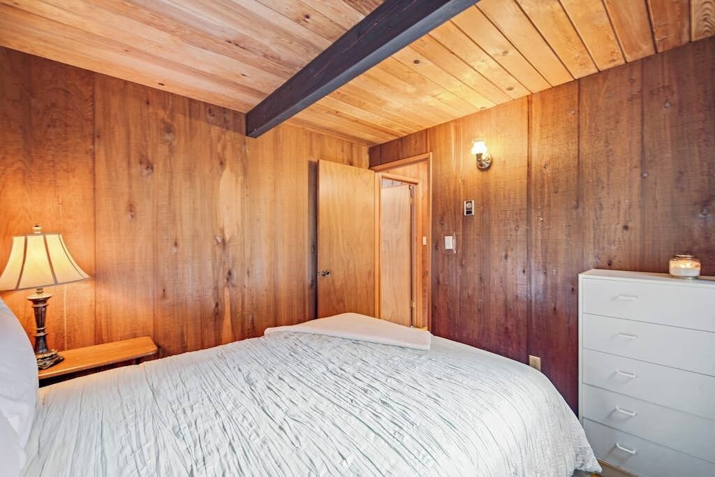 Een kamer met houten wanden, een bed met een witte dekbedovertrek, een bedlampje en een dressoir met lades.