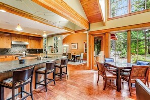 Eine geräumige Küche mit Holzausstattung, einer großen Kochinsel mit Barhocker und einem Essbereich mit Glastisch und Stühlen.