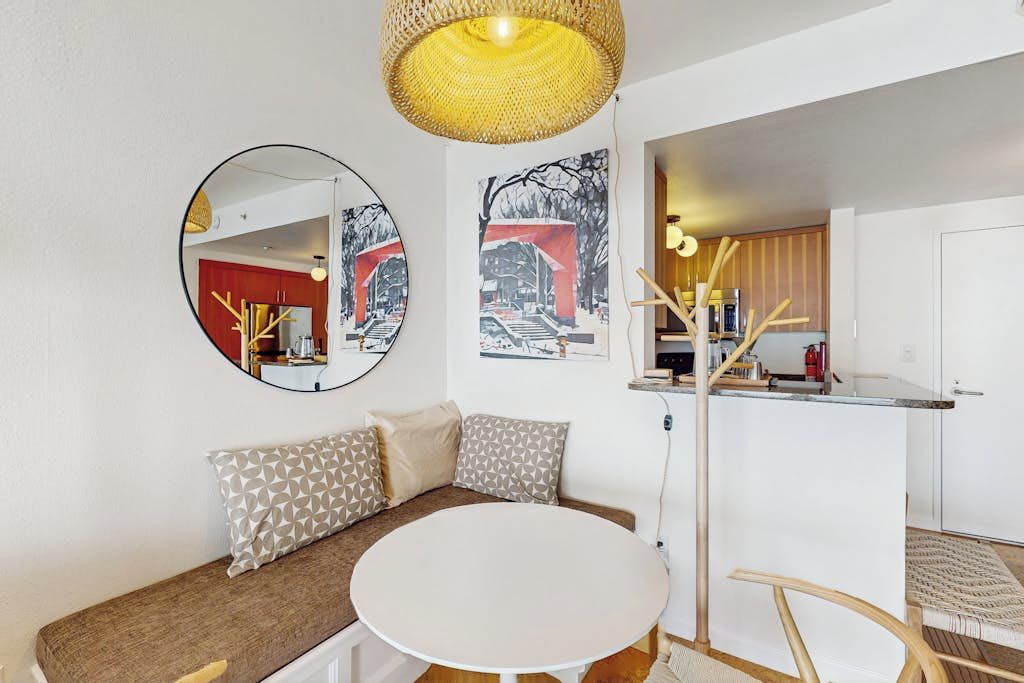 Un espacio habitable moderno con una mesa de comedor redonda, un sofá con cojines estampados y una cocina visible a través de un vano abierto.