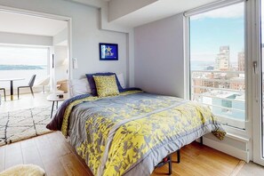 A modern bedroom with a large bed, a small table, and a chair. There is a window with a city view. - 2BR condo with water view, full kitchen, Washer Dryer (Seattle)