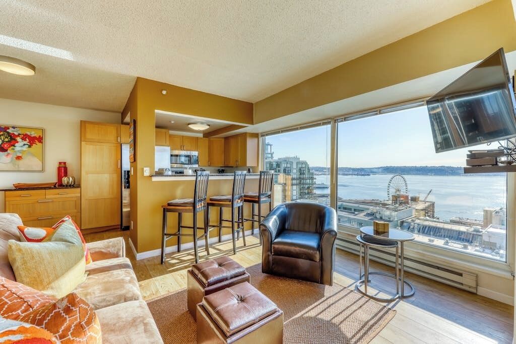 Una sala de estar moderna con un sofá, otomanas, un sillón de cuero y un área de comedor con taburetes altos. Un ventanal ofrece vistas a la ciudad y al agua.
