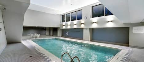 Una piscina interior con suelo de baldosas, una escalera y vistas de la ciudad de noche a través de amplios ventanales.