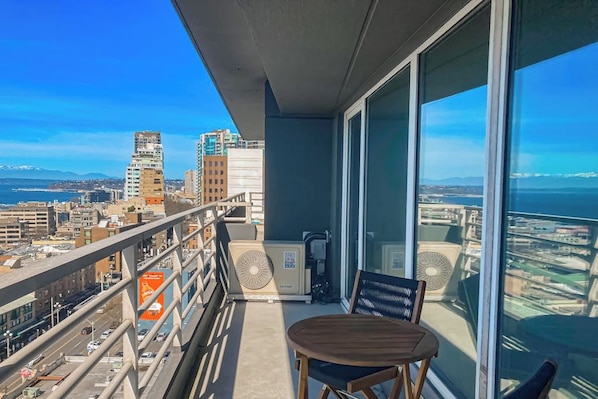A balcony with a table and chairs, overlooking a cityscape with buildings and a body of water. - Luxe 1BR with huge bay view, pool, & hot tub (Seattle)