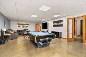 A room with a pool table, a sofa, a television, and a door leading to another room. - 4BR home with pool, hot tub & great location (Tacoma)