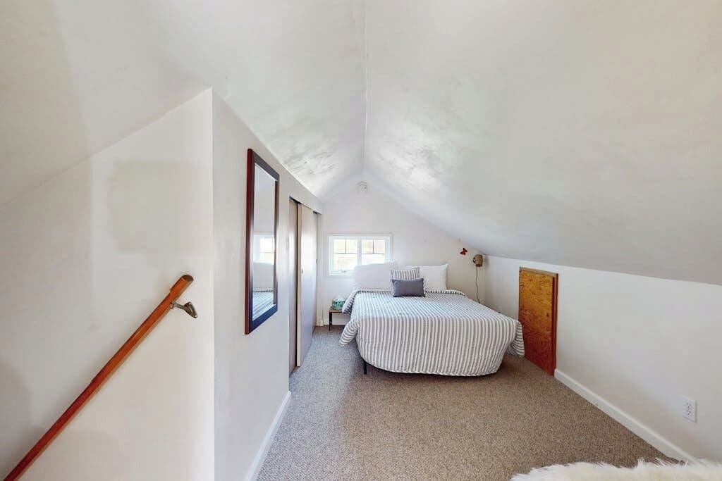 A narrow hallway leading to a bedroom with a sloped ceiling, a bed with striped bedding, and a wooden door.