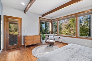 A modern bedroom with a large bed, a wooden dresser, a leather armchair, and a view of trees through large windows. - 4BR mountain-view house withhome office & fireplace (Cle Elum)