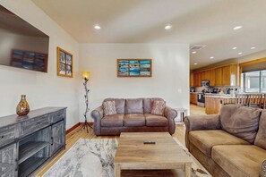 A living room with a sofa, a coffee table, and a TV. There is a kitchen in the background. - 3BR home with hot tub, pool table & wifi (Ronald)