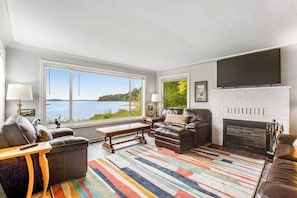 A living room with a fireplace, a large window with a view of the water, and a colorful striped rug.