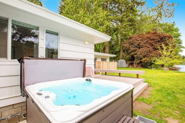 A hot tub outside a house with a wooden deck, a picnic table, and a large tree in the background.