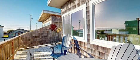 A wooden deck with Adirondack chairs, a wooden wall with a window, and a view of the beach.