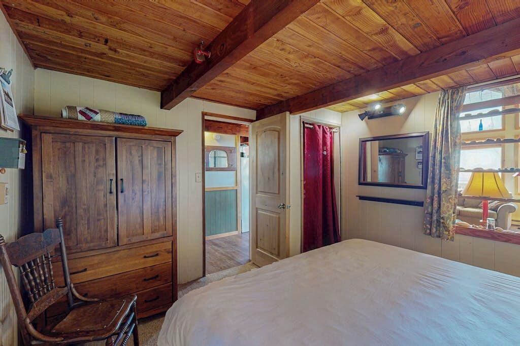 A bedroom with a wooden ceiling, a bed, a wardrobe, a chair, and a window with curtains.