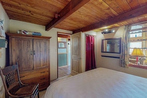 Een slaapkamer met een houten plafond, een bed, een kledingkast, een stoel en een raam met gordijnen.