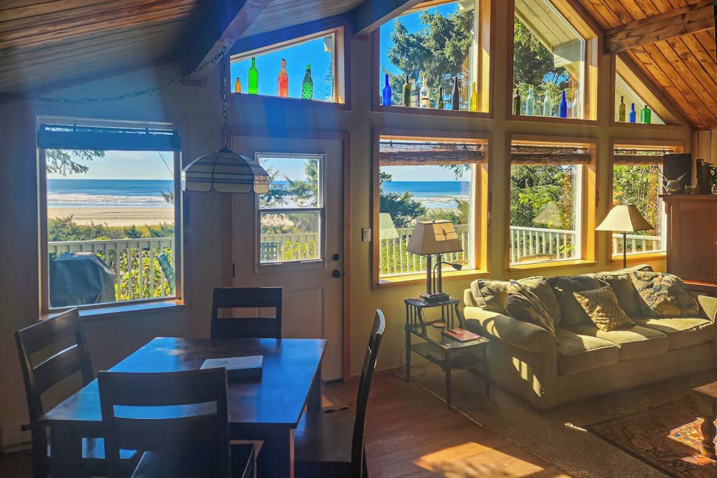A room with a dining table and chairs, a sofa, and large windows with a view of the beach.