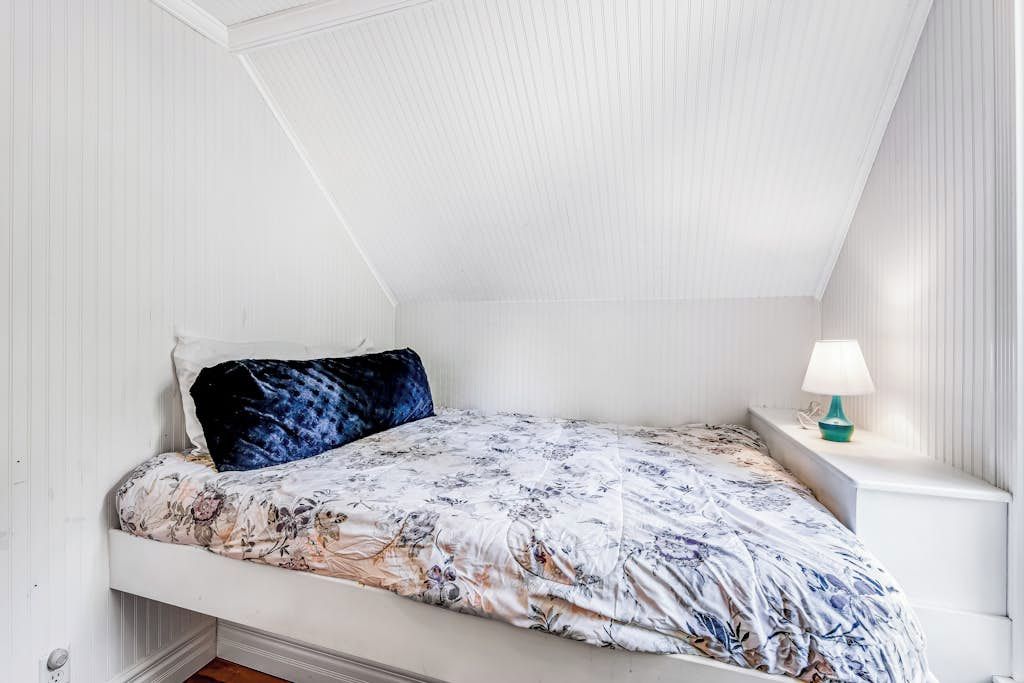 A small, white, single-bed room with a sloped ceiling and a nightstand with a lamp.