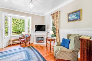 A living room with a fireplace, a television, a patterned armchair, a sofa, and a large window with a view of greenery. - Restored 3BR Dog Friendly - Deck - Firepit - Washer Dryer (Sequim)