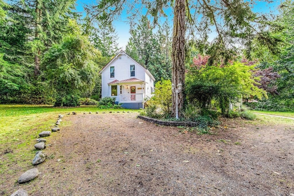 A two-story house with a porch, surrounded by a forested area with a gravel path leading to the entrance.