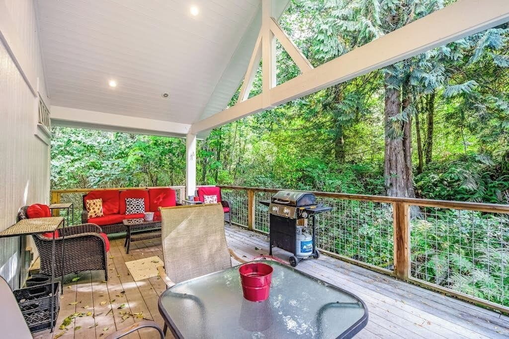 A covered outdoor deck with seating, a grill, and a view of a forest.