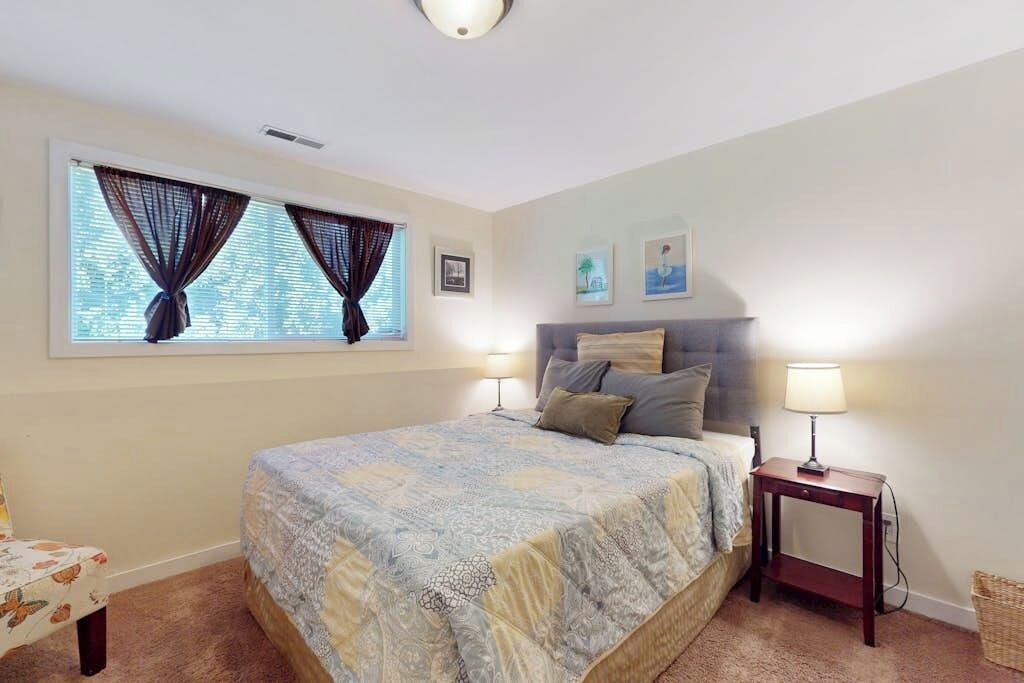 A bedroom with a bed, two bedside lamps, a window with curtains, a small table, and wall art.