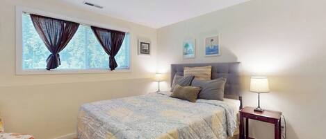 A bedroom with a bed, two bedside lamps, a window with curtains, a small table, and wall art.