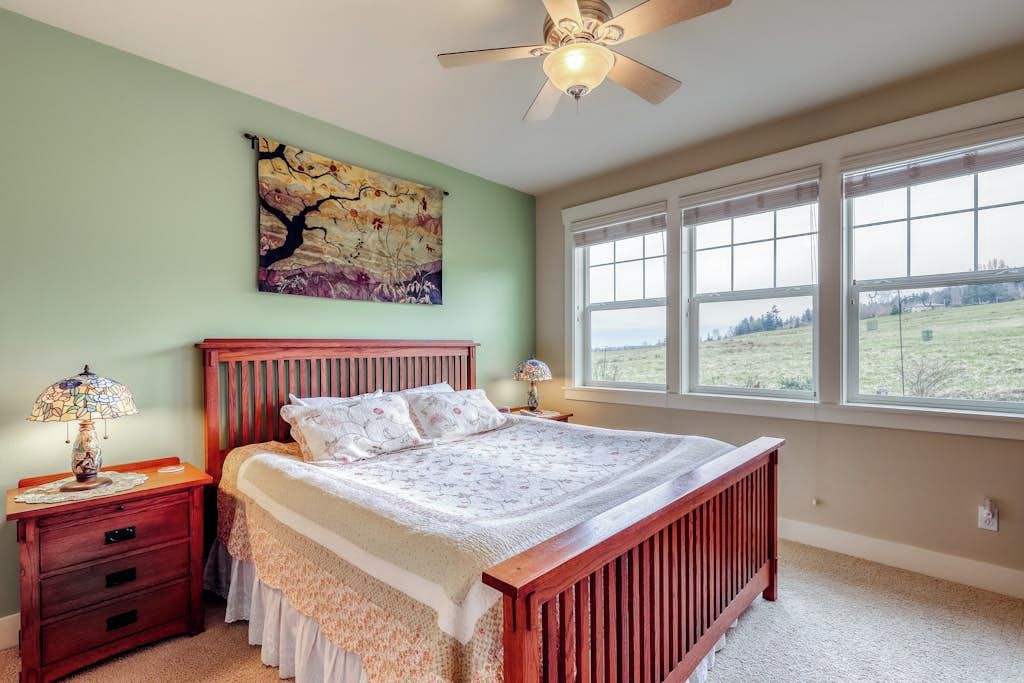 Une chambre à coucher avec un grand lit, une table de chevet, une lampe, un ventilateur de plafond, une fenêtre donnant sur l’extérieur et un tableau accroché au mur.