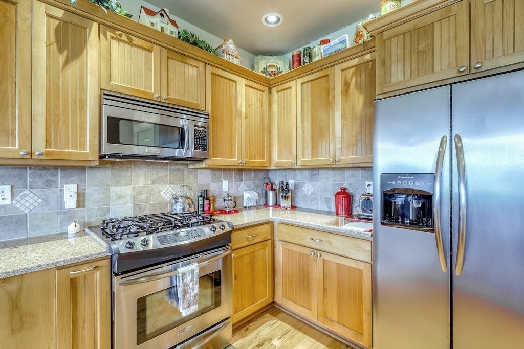 A kitchen with wooden cabinets, stainless steel appliances including a refrigerator, stove, and microwave, and a tiled backsplash.