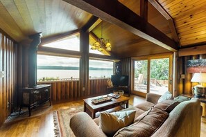 A spacious living room with a large window, wooden furniture, and a television. - 2BR waterfront home with full kitchen & deck views (Poulsbo)