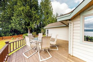 A deck with a table and chairs, surrounded by a wooden deck and a white building with a window. - 2BR waterfront home with full kitchen & deck views (Poulsbo)