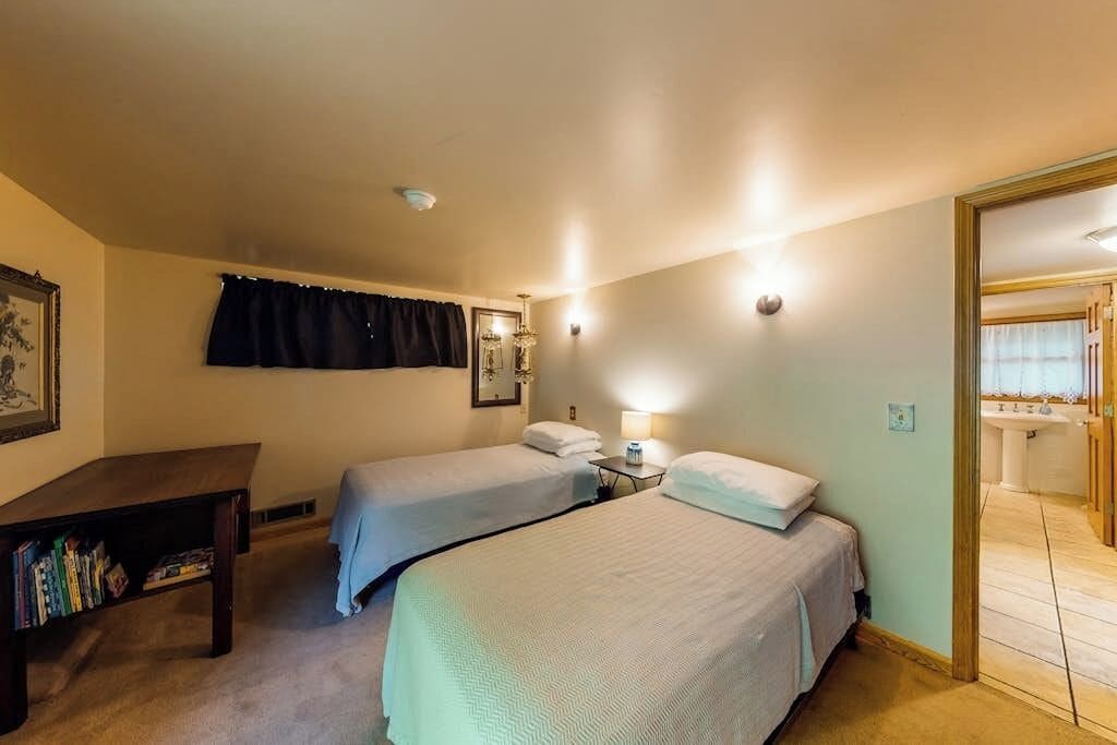 Une chambre d’hôtel avec deux lits, une table de chevet, une lampe, une bibliothèque et une salle de bain visible à travers une porte ouverte.