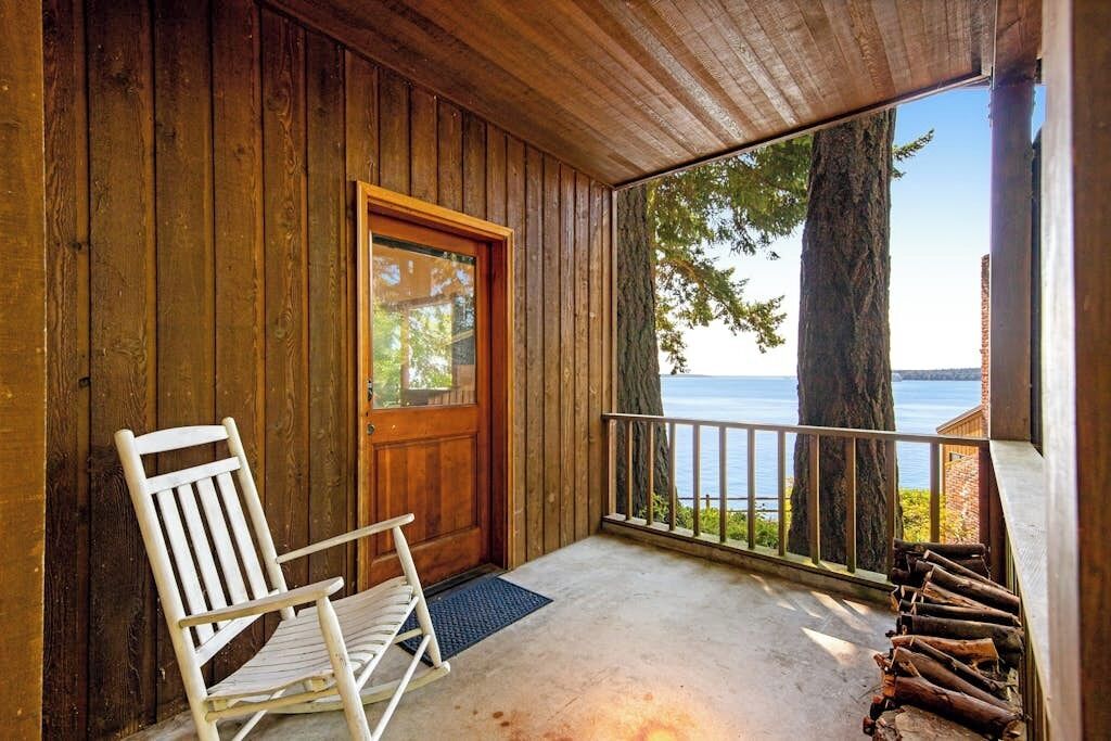 Une véranda en bois avec une chaise berçante blanche, une porte et une vue sur une étendue d’eau.