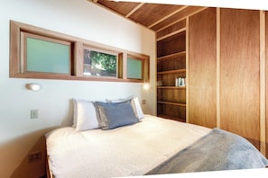 Ein Schlafzimmer mit einem Bett, Kissen, einem Nachttisch, einem an der Wand befestigten Regal mit Büchern und einem Fenster mit Blick auf Bäume.