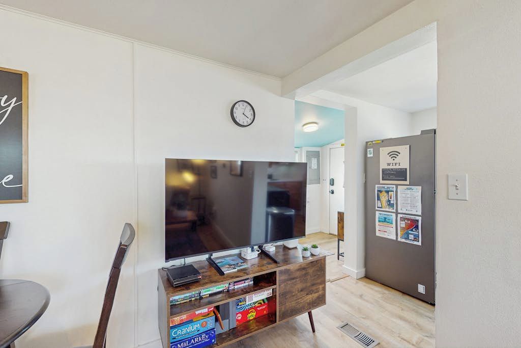 A modern living room with a flat-screen TV on a wooden stand, a dining table with chairs, a clock on the wall, and a refrigerator with a WiFi sign.