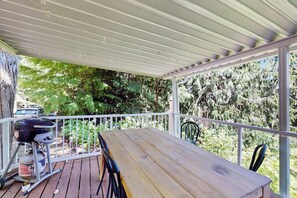 A covered outdoor area with a wooden table and chairs, a grill, and a view of trees.