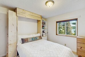 A bedroom with a bed, a window with a view, a wooden shelf unit, and a ceiling light.