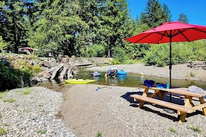 A riverside picnic area with a red umbrella, wooden benches, and inflatable rafts. - 4BR luxe lodge with hot tubs, kayaks, & firepits (Tahuya)