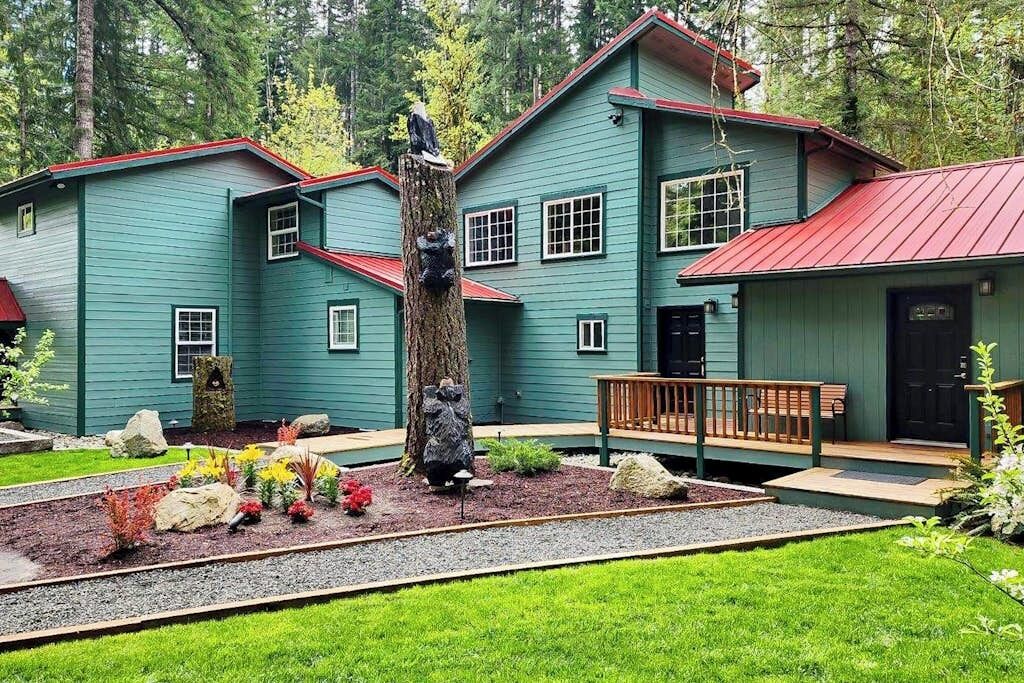 Una casa de dos pisos con techo rojo y revestimiento verde, rodeada por un jardín ajardinado con diversas plantas y una terraza de madera.