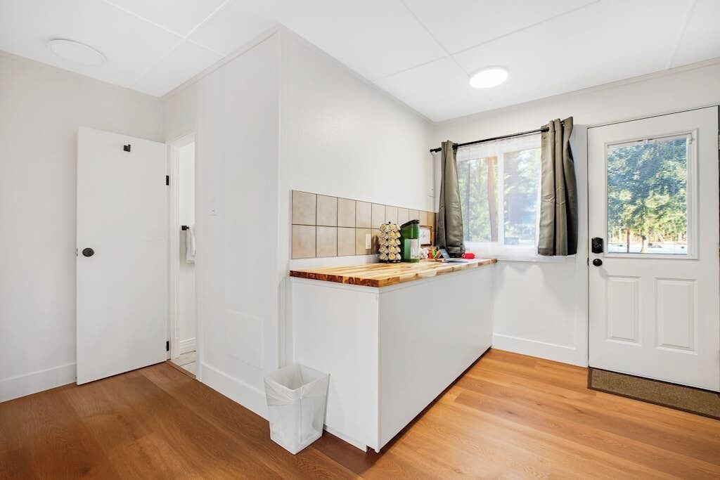 Quarto com piso de madeira, paredes brancas, painel de azulejos, janela com cortinas e uma porta.